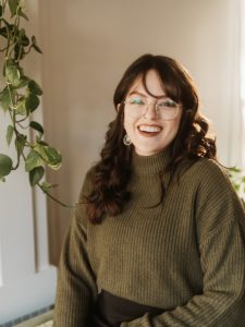 A picture of Adina in a denim shirt and a grey beanie sitting against a white wall with black symbols drawn on it. These include the Scorpio glyph, a daisy, a bone, a fish, and a ufo.